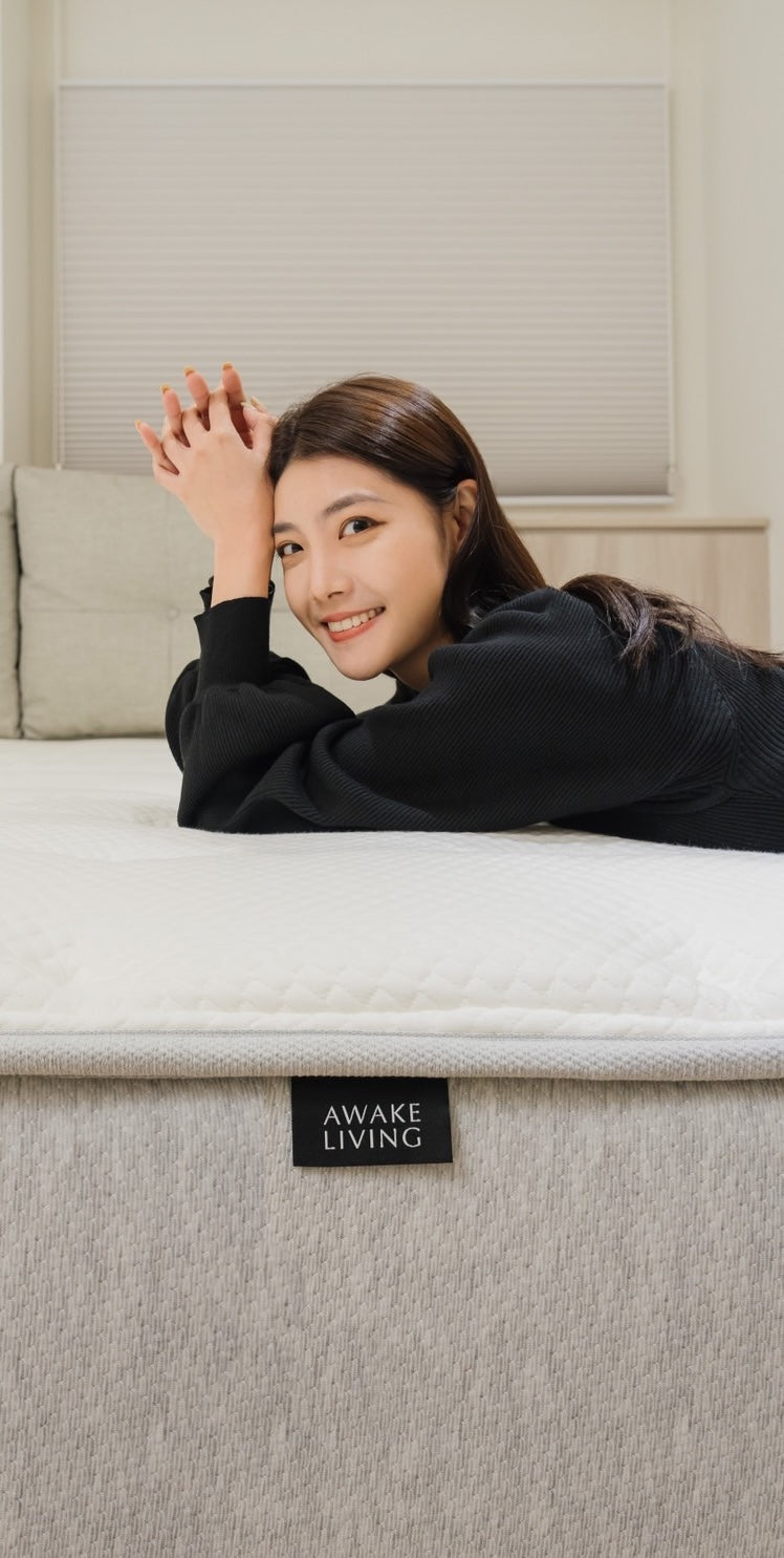 一位面帶微笑的女性趴在床墊上，前景清楚地顯示 Awake Living 品牌布標。