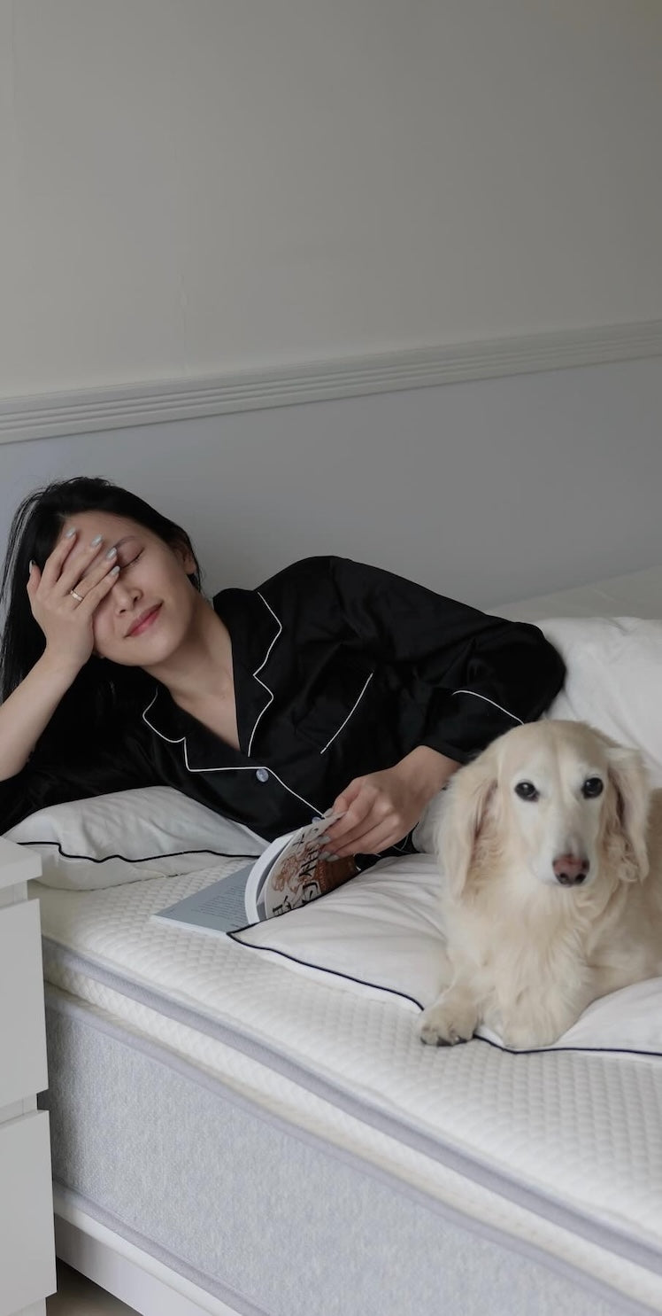 一位閉眼手撫額頭的女性，與一隻臘腸狗一同躺在床上休息。