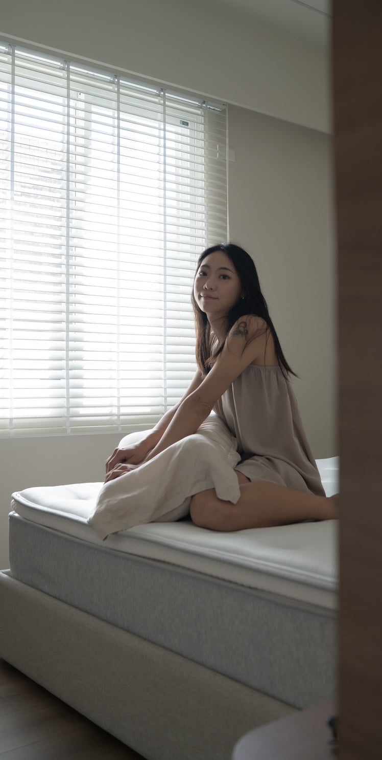 一位女性坐在床沿，身後是透著光的白色百葉窗，前景為門框。