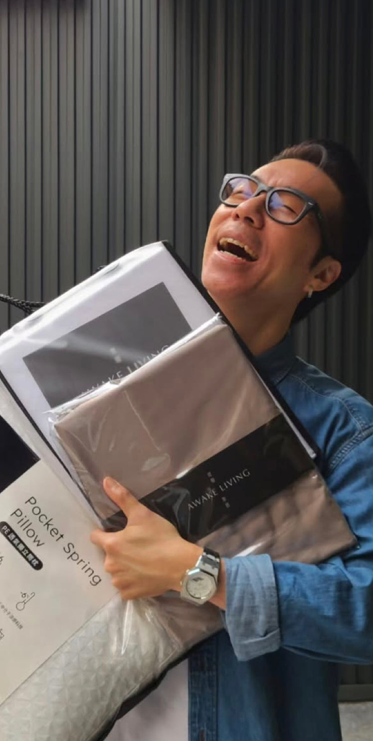 一位戴著眼鏡的男子開懷大笑，懷裡抱著一疊 Awake Living 的枕頭與寢具產品。