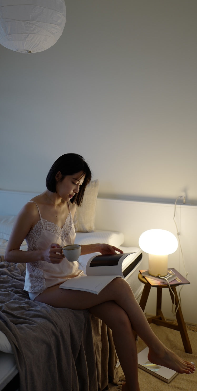 一位女性坐在床上，藉著床邊檯燈的燈光閱讀書本，並手持馬克杯。