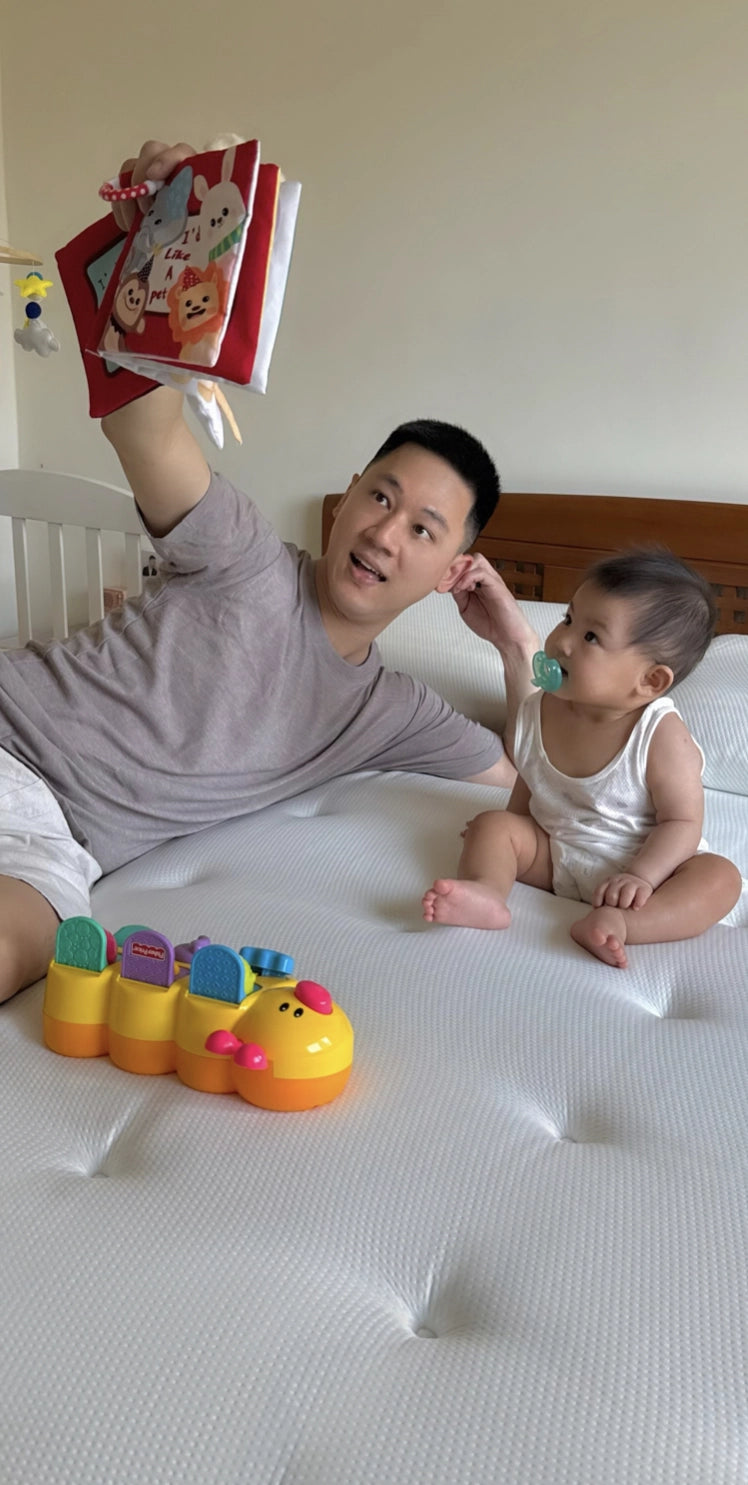 一名男子與嬰幼兒一同躺在白色的床墊上，手持繪本念著故事給幼兒聽
