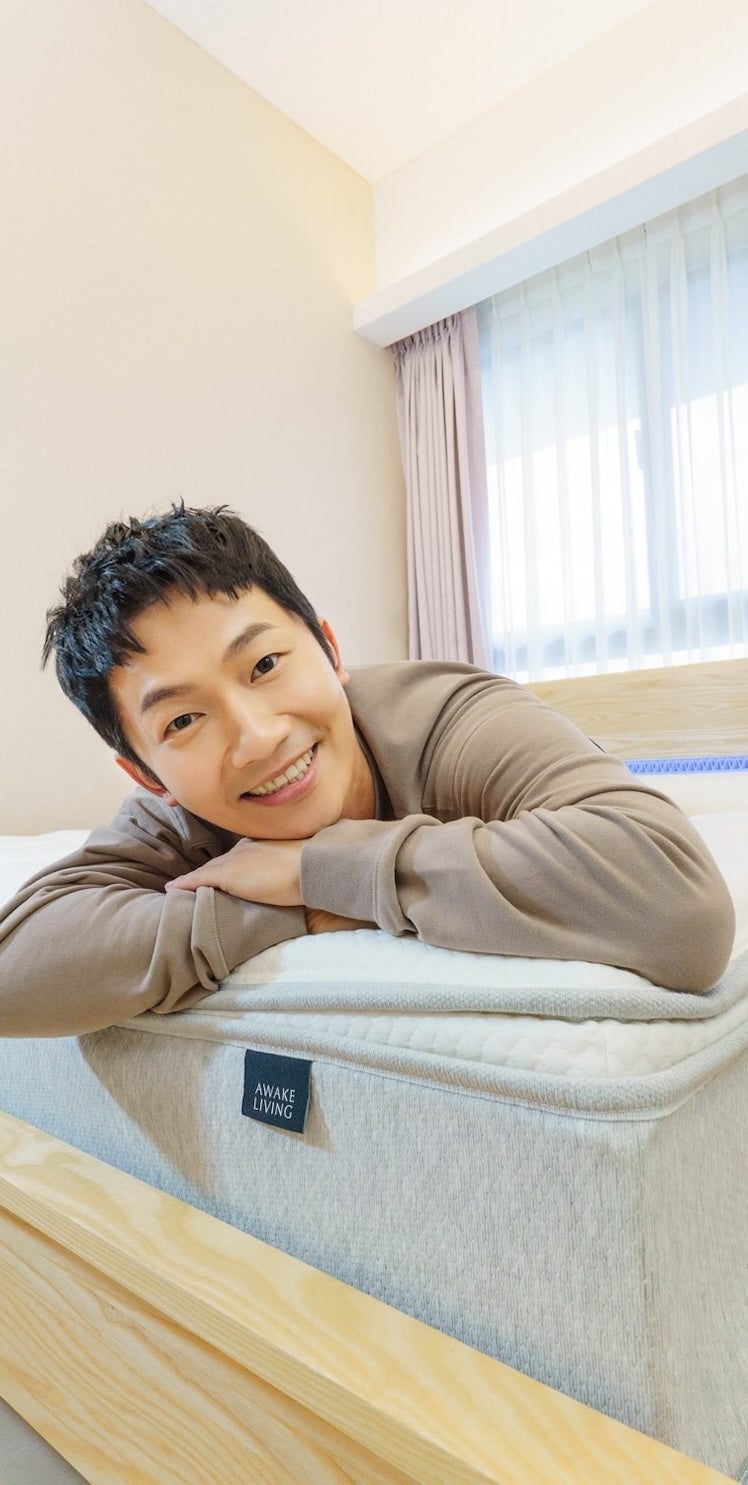 一位面帶微笑的男性趴在床墊上，前景清楚地顯示 Awake Living 品牌布標。